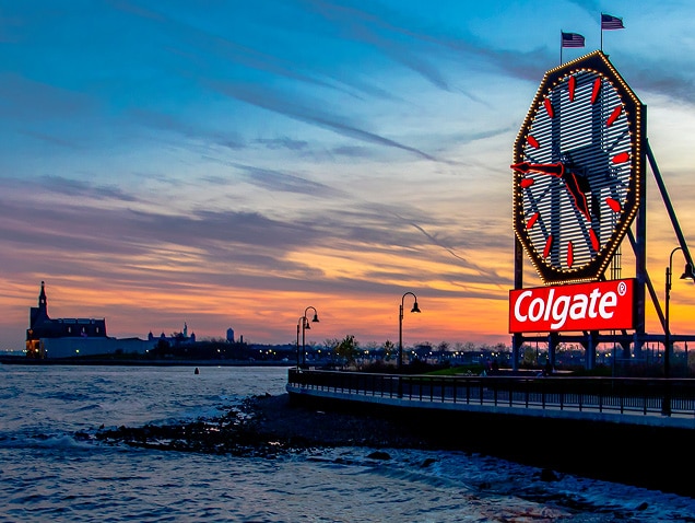 jūras krasts, Colgate logotips fonā zem liela pulksteņa ar sarkaniem rādītājiem