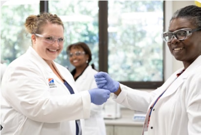 Divi zinātnieki laboratorijas halātos un cimdos smaidot spiež rokas laboratorijas vidē
